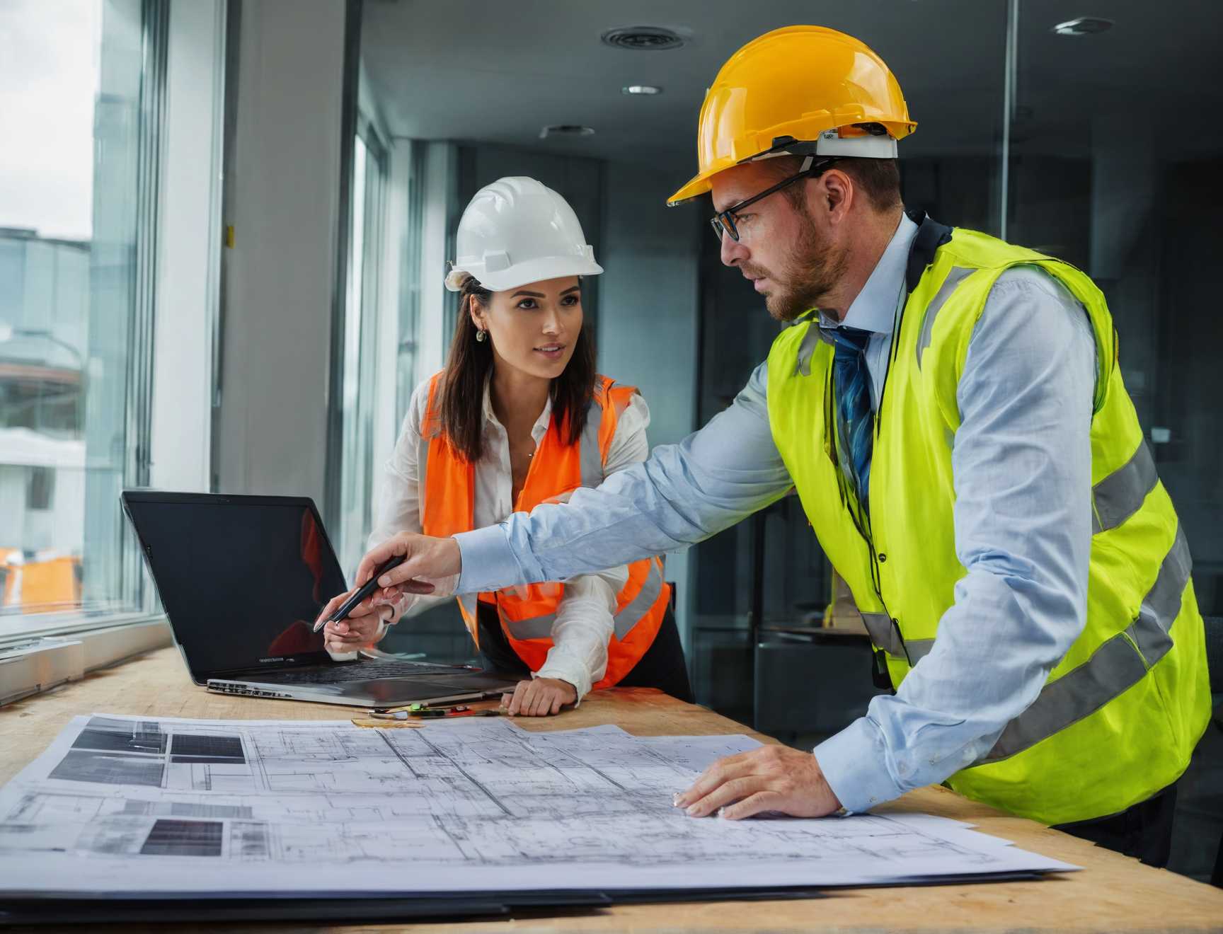 Deux personnes en casque et gilet de chantier étudient des plans devant un ordinateur portable